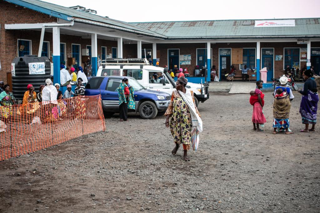 People walk and gather outside a building with a rusted roof in a rural setting. Some individuals wear colorful, traditional clothing. A vehicle is parked nearby, and there is an orange plastic barrier separating certain areas. The atmosphere appears busy.