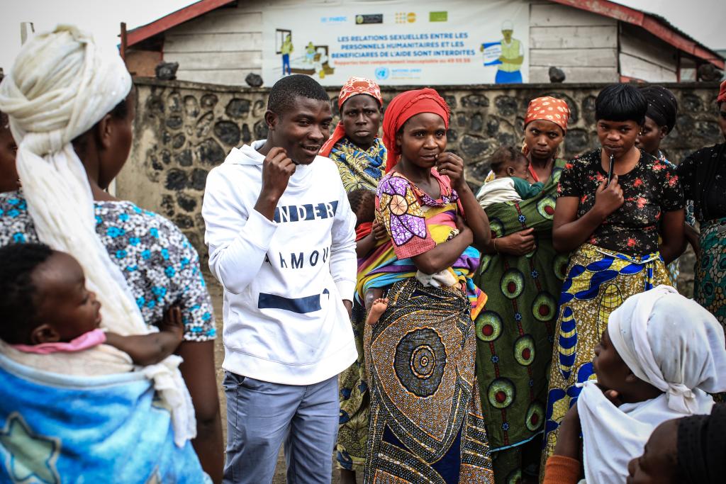 A young man in a white sweatshirt leads a discussion with a group of women, some with babies, who are dressed in colorful patterns. They are standing outdoors with a building in the background displaying text in French. The atmosphere appears engaged and attentive.