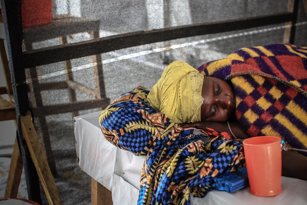 A person rests on a bed covered with vibrant patterned blankets and a yellow headscarf. An orange cup sits nearby on the bed. The background is minimal, with a mesh barrier visible.