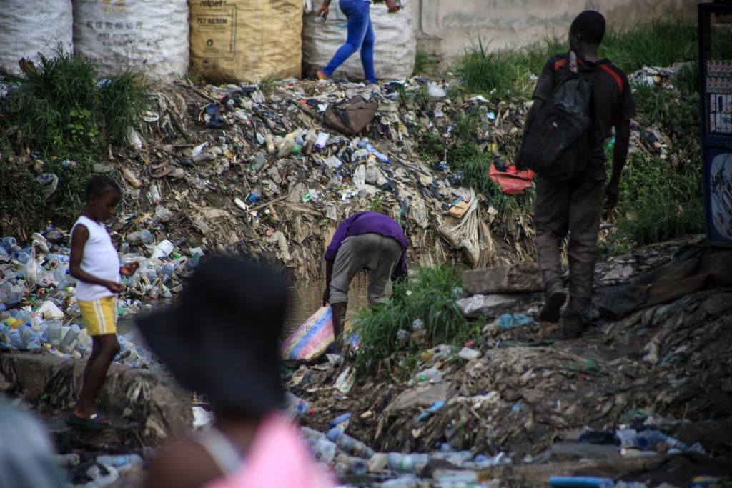 People are foraging through a large area filled with plastic waste and debris. One person is bending down, picking up something from the ground, while others are walking around the area. Large bags filled with waste are visible in the background.