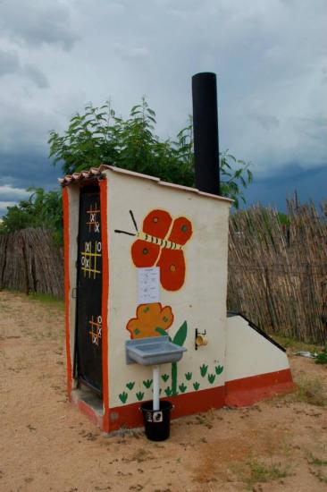 A small outdoor structure with white walls and a red border, decorated with a large painted butterfly and simple geometric designs. A faucet with a small sink and bucket is mounted on the side. It stands in a sandy area with a cloudy sky overhead, surrounded by a wooden fence.