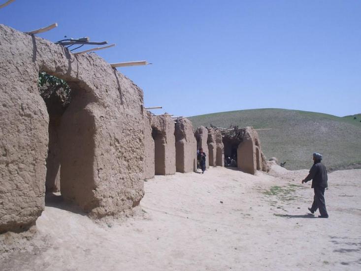 A person stands near a row of mud-brick structures with arched doorways in a semi-arid landscape. Wooden beams protrude from the tops of the structures. The background features a clear blue sky and rolling hills with sparse vegetation.