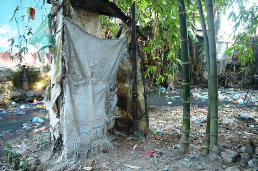 A makeshift structure made of tattered cloth stands beside a polluted stream amid bamboo trees. Trash littering the ground contrasts with the natural surroundings, highlighting environmental and living condition issues.