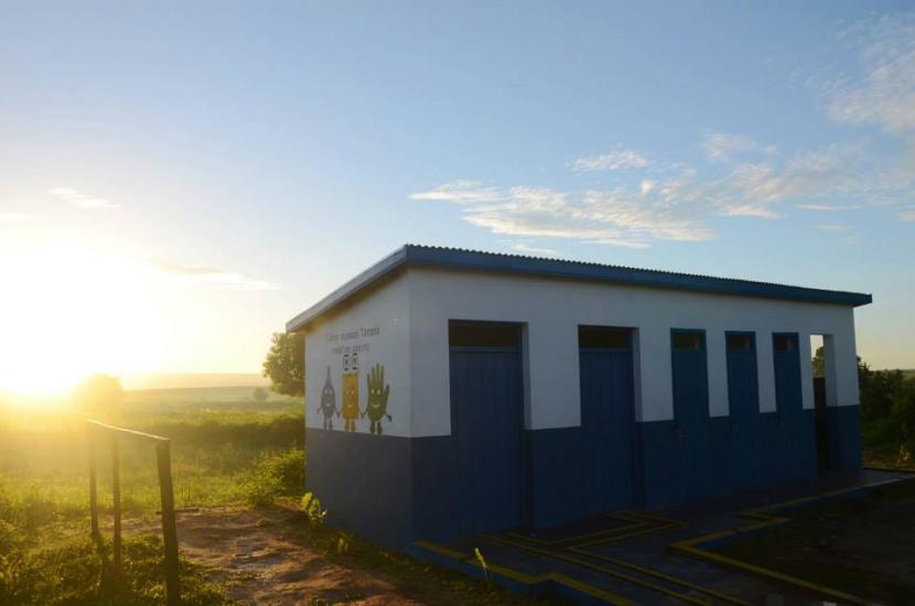 A small white and blue building with multiple doors stands in a rural area. The sun is setting, casting a warm glow over the landscape. Simple cartoon characters are painted on the building's wall. Lush greenery surrounds the structure.