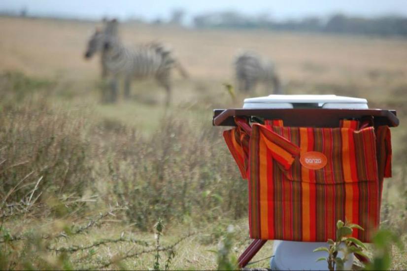 A portable toilet with a striped red and orange bag hangs on its side, sits in a grassy field. Two zebras graze in the blurry background, showcasing a savanna landscape. The sky is overcast with soft light. .