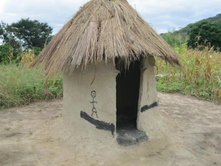 A small, round mud hut with a thatched roof stands in a rural area. The hut has an open doorway and simple stick-figure drawings on its walls. Surrounding the hut are green plants and trees, and the ground is bare with patches of dirt.