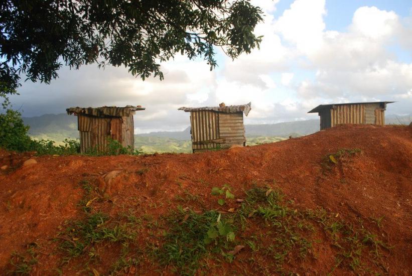 Three small, rustic wooden sheds are situated on a reddish, elevated terrain with green foliage at the foreground. The backdrop features a scenic view of rolling hills and clouds under a partly cloudy sky, with a large tree branch extending from the top left corner.