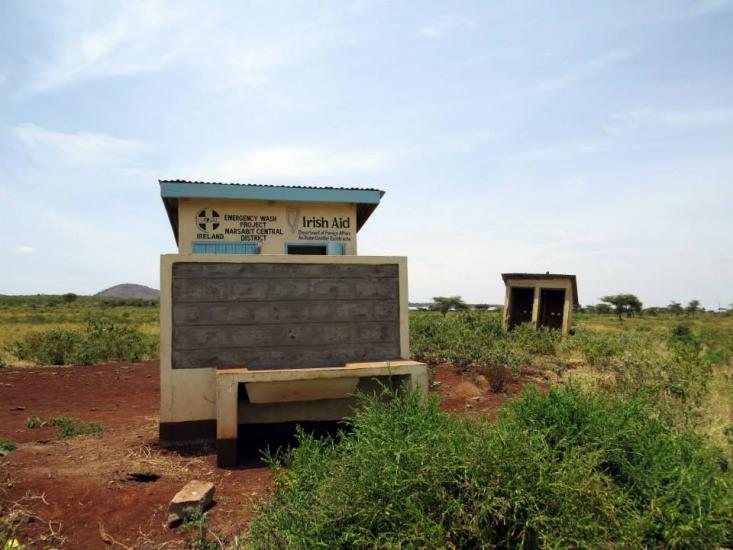 A small, rural medical facility is shown with a concrete structure displaying a sign mentioning Irish Aid support. The area surrounding the facility is grassy and expansive, with a few scattered trees and a small outbuilding in the background. The sky is clear and blue.