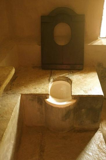 A rustic, historical toilet made of stone and wood. The toilet has a wooden seat with a round opening and a stone base. The setting appears to be dimly lit, with light coming in from a small window to the right. The design looks ancient and well-worn.