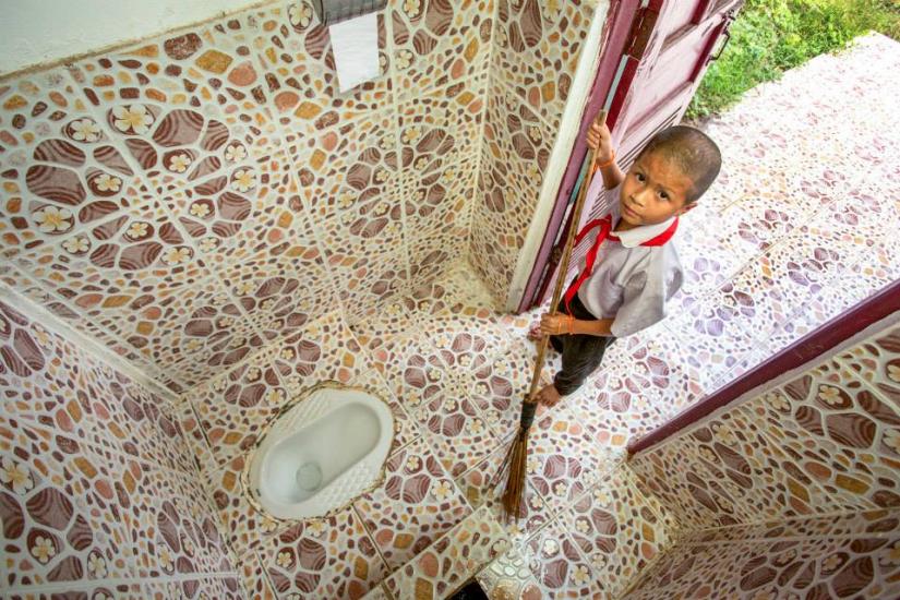 A young boy in a school uniform stands in the doorway of a small bathroom with mosaic-patterned tiles. The bathroom features a squat toilet and a roll of toilet paper mounted on the wall. The door opens to a tiled pathway leading outside.