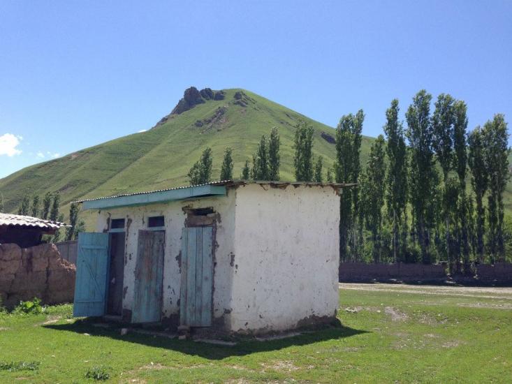 A small, weathered white building with blue doors stands in front of a lush, green hill. Tall, thin trees line the background under a clear blue sky.