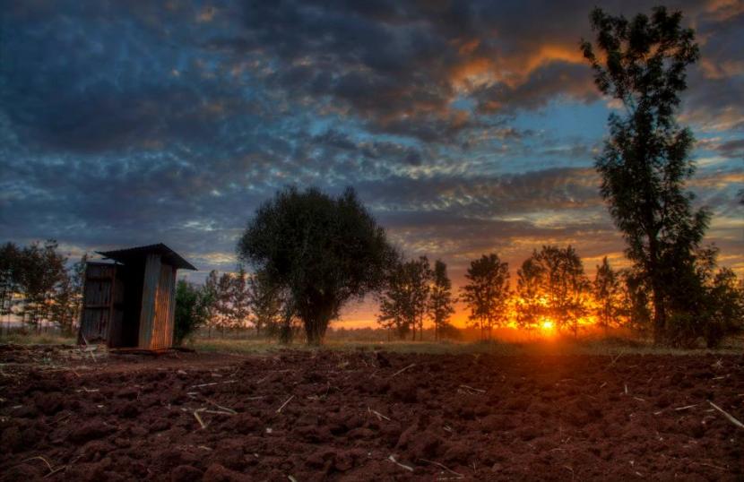 A rustic shed stands in an open field at sunrise, with a dramatic sky of blue and orange hues. The ground is plowed, and trees surround the area, creating a serene rural landscape.