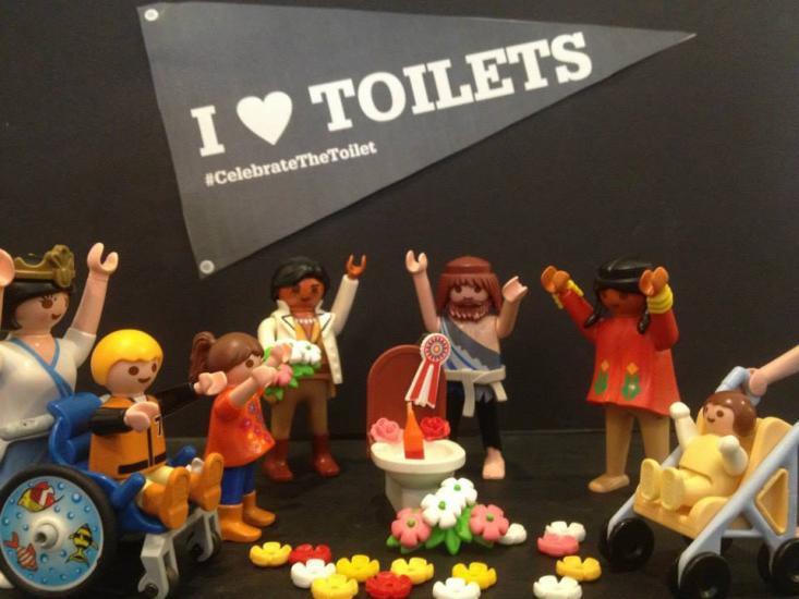 Toy figurines celebrate around a decorated toilet with a banner reading "I ♥ TOILETS #CelebrateTheToilet" in the background. Some figures are in wheelchairs and others are on foot, surrounded by colorful flowers on the ground.