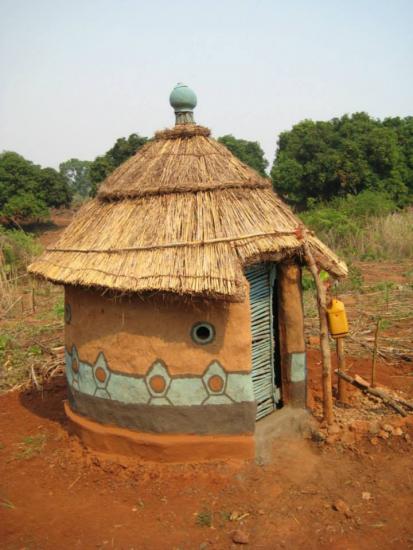 A small circular hut with a thatched roof and geometric painted designs on its mud walls stands on red soil. It has a closed metal door and a rounded window. The surrounding area is a mix of greenery and dry patches of land.