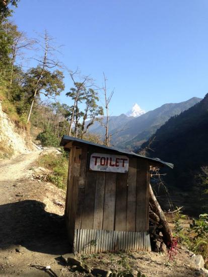 A small wooden toilet is situated alongside a dirt path in a mountainous area. Trees are scattered around, and a distant peak is visible against a clear blue sky.