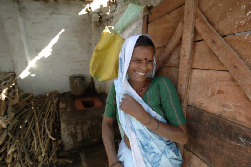 A woman wearing a green blouse and a white sari with a blue pattern stands smiling against a wooden wall. She has a yellow bag slung over her shoulder and is in a rustic, dimly lit setting with some sticks and a container visible in the background.