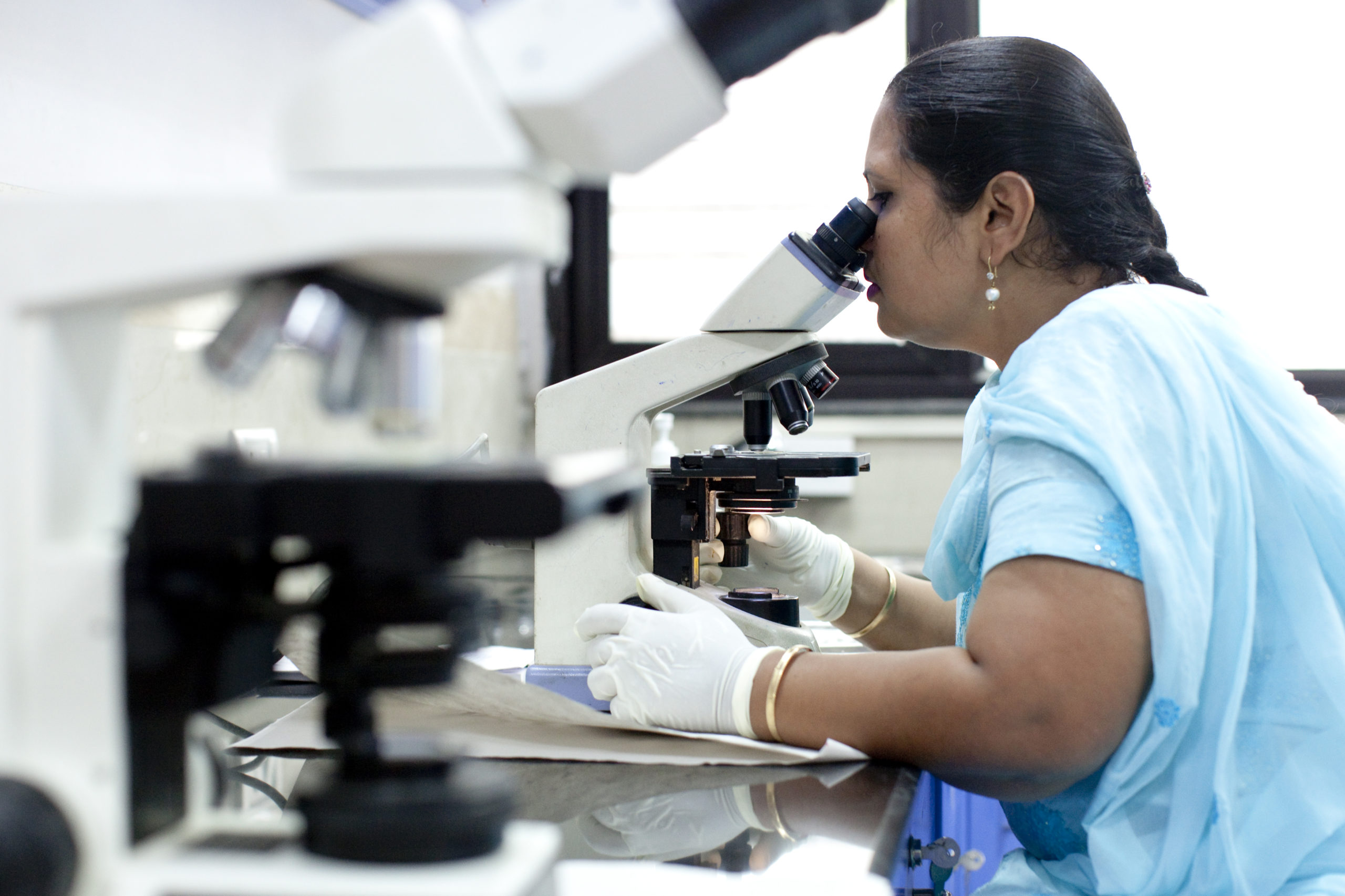 A woman in a blue outfit and gloves looks through a microscope in a lab. Another microscope is visible in the foreground. She appears focused on her work, seated at a lab bench.