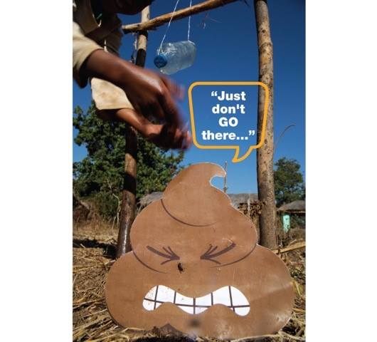 An outdoor setup with a plywood cutout of a poop emoji with an angry face. A hand is seen adjusting a hanging plastic bottle above it. A speech bubble next to the cutout states, "Just don't GO there..." The background shows a clear sky and sparse greenery.