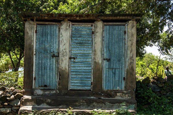 A weathered concrete structure with three rusted metal doors, set against a lush, green backdrop with trees and shrubs. The structure appears to be old and run-down, and there are some scattered stones and a person partially visible in the distance on the right.