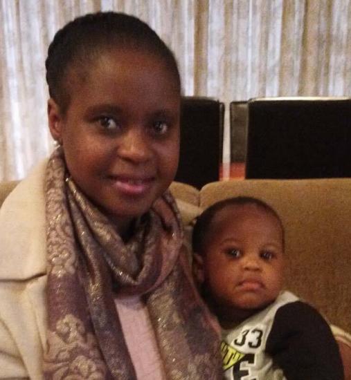 A woman with a patterned scarf around her neck holds an infant on a sofa. They both face the camera, and a curtain is in the background. The baby is wearing a black and gray shirt.