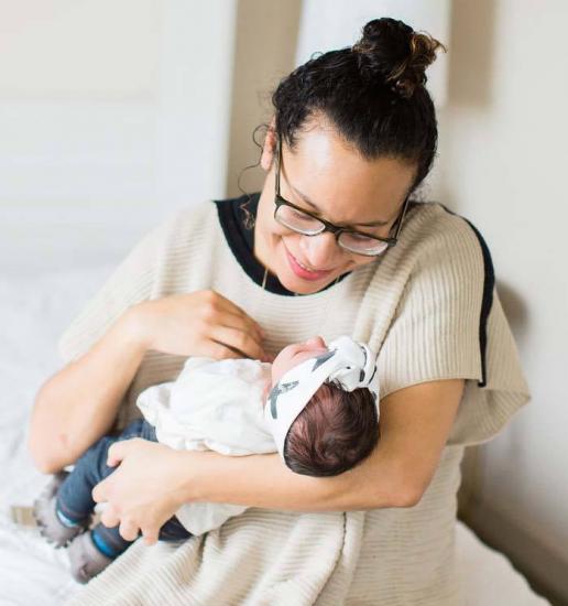 A person wearing glasses and a beige top is sitting on a bed and smiling while holding an infant dressed in white with a white headband. The infant appears to be asleep or calm in their arms. The setting is a cozy, softly lit room.