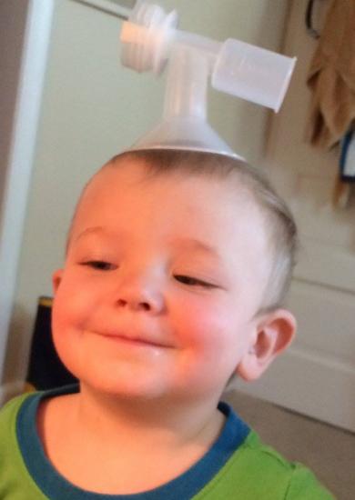 A young child with a mischievous smile has placed a funnel-like object on their head like a hat. The child appears amused and is wearing a green shirt with blue trim. The background shows an interior space with a door and some hanging items.