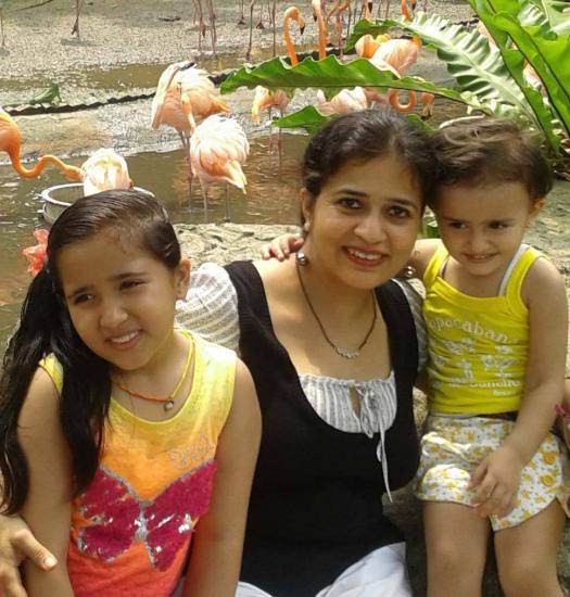 A woman sits outdoors with two young children at a zoo. They are smiling and appear happy. Behind them, pink flamingos are standing and wading in a pond. Lush greenery surrounds the area.