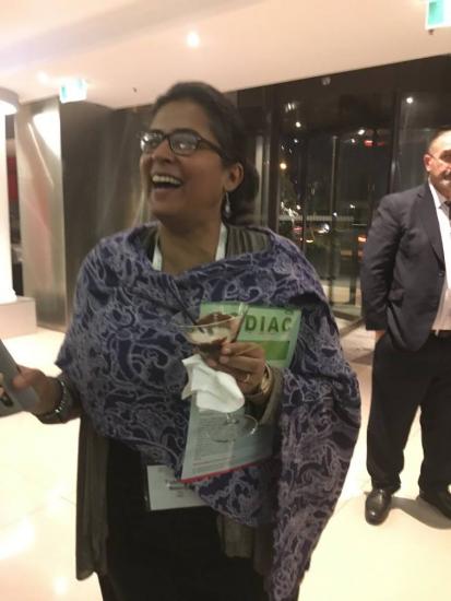 A woman is joyfully smiling and holding a cocktail in one hand and a phone in the other. She is wearing glasses, a patterned scarf, and has a conference badge around her neck. A man in a suit stands in the background within a well-lit indoor setting.