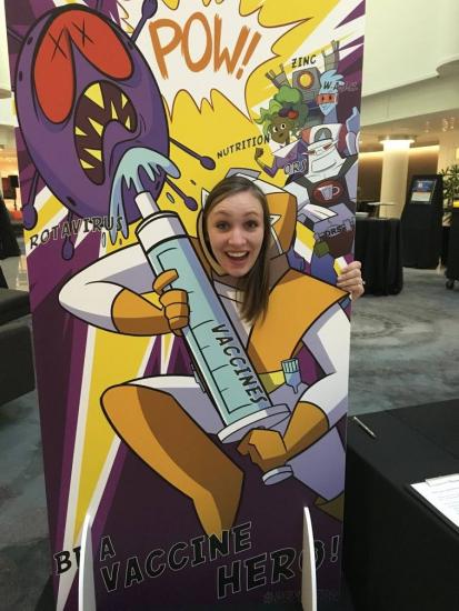 A woman joyfully poses behind a cutout stand featuring an illustrated superhero-themed vaccine poster. The colorful poster shows the superhero fighting off cartoon viruses with a large syringe labeled "vaccine," accompanied by action words like "POW!.