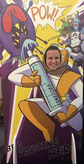 A person poses with their face in a cutout of a cartoon astronaut holding a large syringe labeled "VACCINE" aimed at a purple virus, with comic-style elements such as "POW!" and "Be a Vaccine Hero!" in the background. Other cartoon figures promote nutrition and zinc.