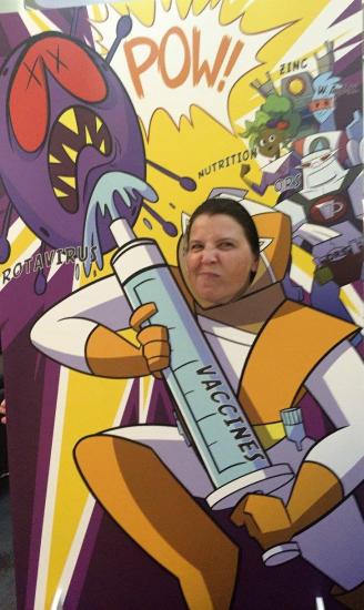 A person poses with their face in a cutout of a cartoon superhero holding a giant syringe labeled "VACCINES" aimed at an anthropomorphized virus. Bright comic-style graphics include "POW!" and words like "NUTRITION" and "ZINC" in the background.