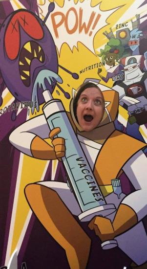 A person playfully sticks their face through a cutout of an illustrated scientist holding an oversized syringe labeled "VACCINE" while battling a cartoon representation of a virus. The background is colorful with comic-style effects and text.