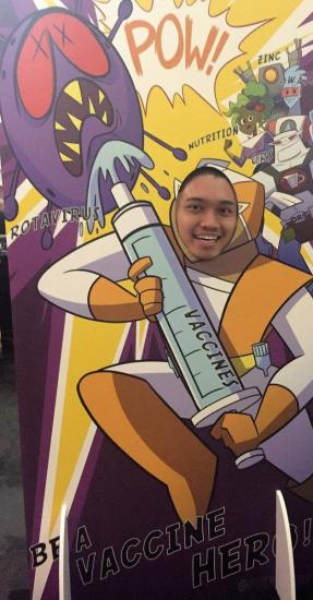 A person smiling through a cutout of a cartoon superhero holding a large syringe labeled "Vaccine." The backdrop includes the words "Be a vaccine hero!" and features stylized comic elements and a menacing cartoon virus.