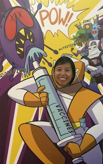 A person playfully poses with their face in a cartoon cutout, showing a superhero figure holding a large syringe labeled "VACCINES," battling a purple virus. Sound effects like "POW!" are illustrated in the background.