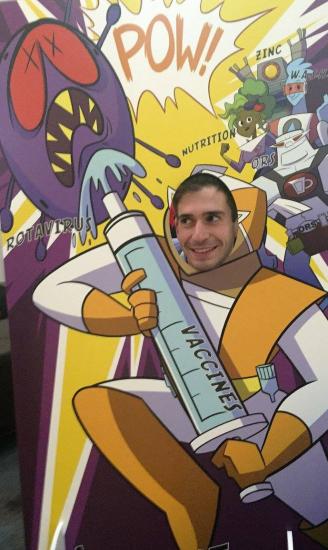 A person poses with their face through a cutout in a cartoon display. The display features a superhero injecting a large syringe labeled "VACCINES" into a monstrous virus. The scene includes text like "POW!" and labels such as "ROTAVIRUS," "NUTRITION," and "ZINC.