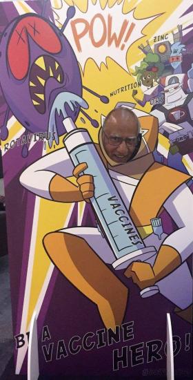 A person poses with their face in a cut-out board designed like a comic-book superhero scene. The superhero holds a large syringe labeled "VACCINE," targeting a menacing virus creature. The background features action words like "POW!" and "ZING!" along with the caption "BE A VACCINE HERO!.