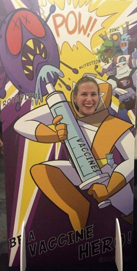 A person poses with their face inserted into a colorful cutout of a superhero holding a giant syringe labeled "VACCINE" facing a purple monster. The background features comic-style text including "POW!" and promotes being a vaccine hero.