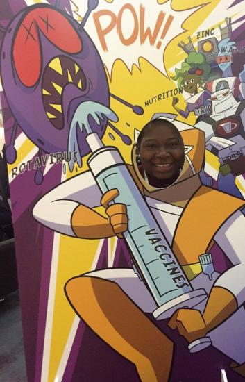 A person smiling through a cutout in a colorful cartoon display. The display depicts a superhero holding a giant syringe labeled "Vaccines" aimed at a purple virus labeled "Rotavirus." Comic-style text and illustrations are in the background.