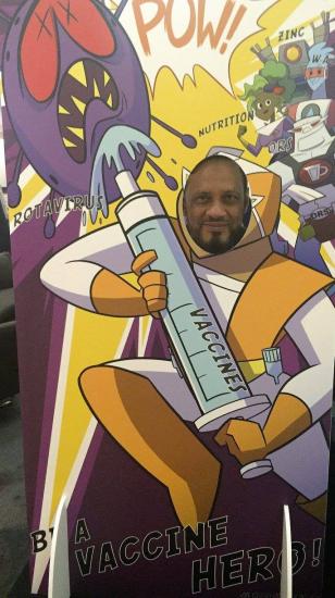 A person poses behind a cartoon cutout featuring a superhero holding a large syringe labeled "vaccine." The background includes comic-style text and illustrations, with phrases like "Pow," "Rotavirus," "nutrition," and "zinc." The scene promotes becoming a "vaccine hero.