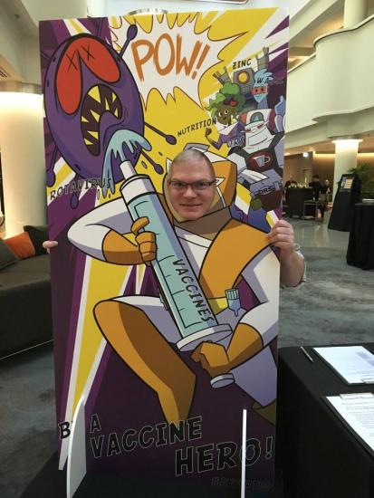 A person poses with their face in a comic-style standee featuring a superhero character holding a large vaccine syringe. The background includes colorful words like "POW!" and illustrations of viruses being defeated. Text reads "Be a Vaccine Hero!.