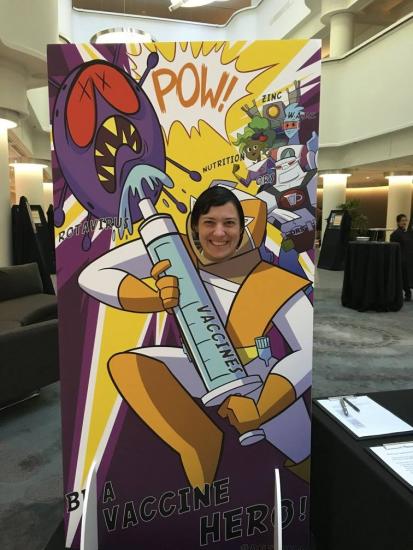 A person stands behind a cutout board featuring a comic-style superhero holding a giant syringe labeled "Vaccine." The superhero is fighting a purple virus character. Words like "POW!" and "BLAM!" are illustrated in the background, along with depictions of vitamins.