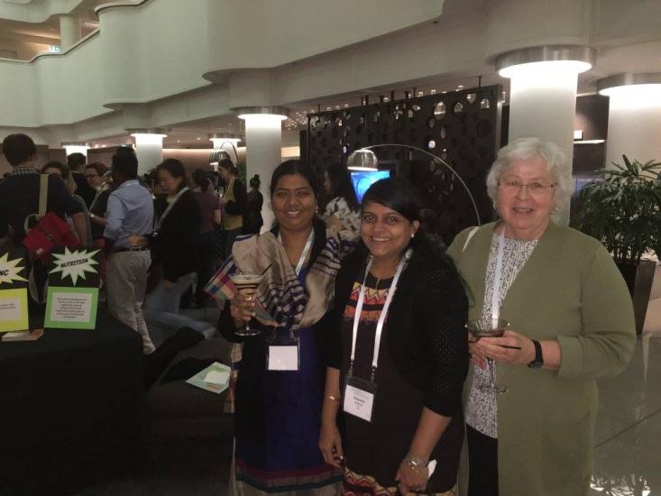 Three women are smiling and standing together at an indoor event. The first two women wear traditional Indian attire, and the third woman is in a green sweater. They are holding drinks and wearing name badges around their necks. Other people and displays are in the background.