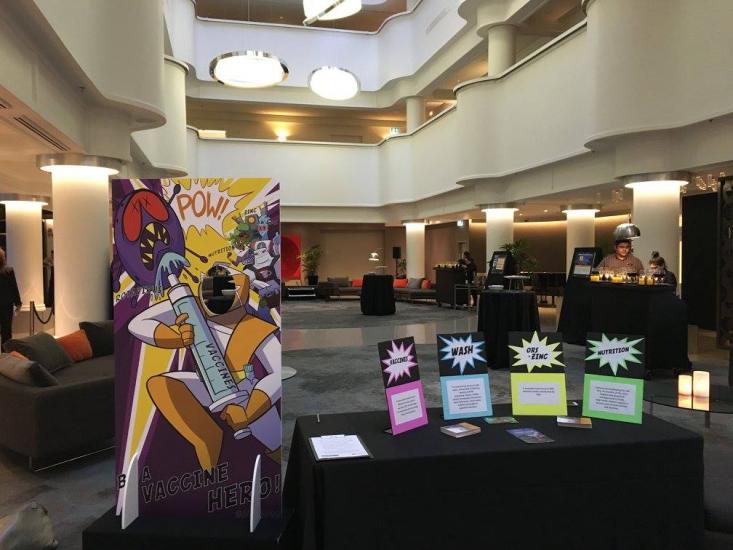 A spacious conference hall with a superhero-themed vaccination cutout stand, tables with informational signs about health safety (mask, distance, sanitize), and people engaging in conversations. The hall has modern lighting and plush seating arrangements.