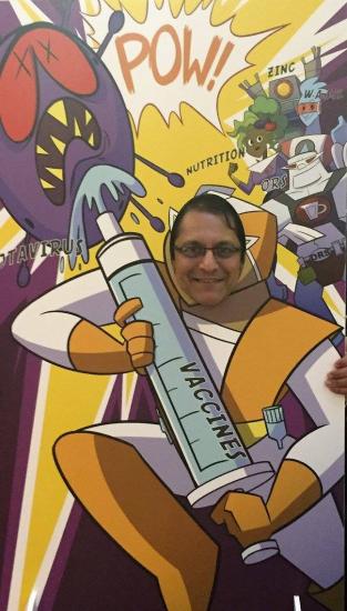 A person poses with their face in a comic-style cutout depicting a superhero holding a large syringe labeled "VACCINE" aimed at a menacing virus. The background features sound effects like "POW!" and elements like "ZINC" and "NUTRITION," symbolizing fighting disease.