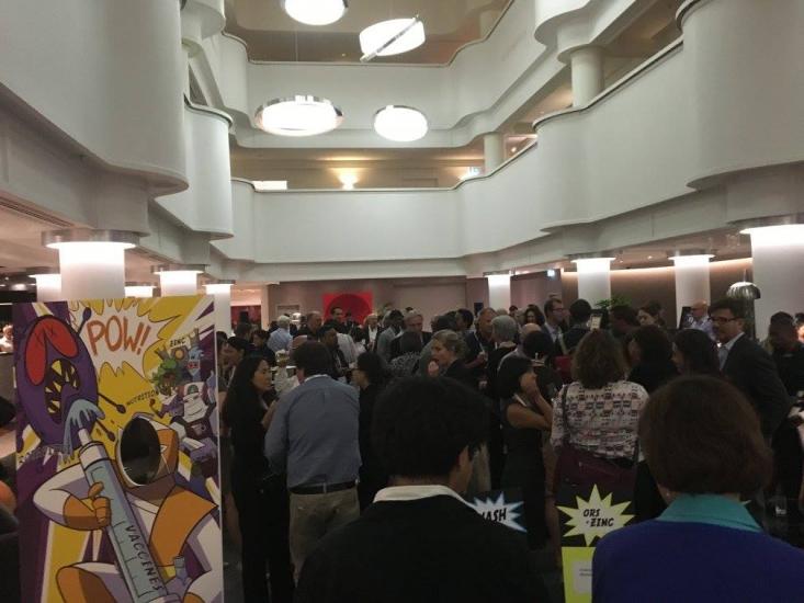 A crowded indoor event with many people milling about and conversing. The room has layered balconies with modern, round ceiling lights. In the foreground, there are colorful comic-style decorations with illustrations and speech bubbles like "POW!" and "CRASH!".