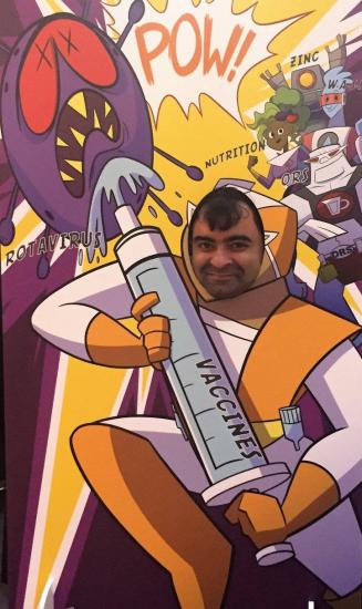 A person poses with their face in a cutout of a cartoon superhero holding a giant syringe labeled "VACCINES." The superhero is defeating a purple villain labeled "ROTAVIRUS," with a comic-style "POW!" The background includes items labeled "NUTRITION," "ZINC," and "ORS.