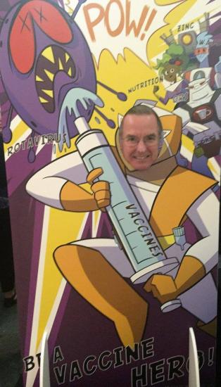 A man poses with his face in a cutout of a cartoon superhero holding a large vaccine syringe. The backdrop is vibrant, with illustrations of various elements like viruses and the words "POW!" and "Be a Vaccine Hero!" in bold text.