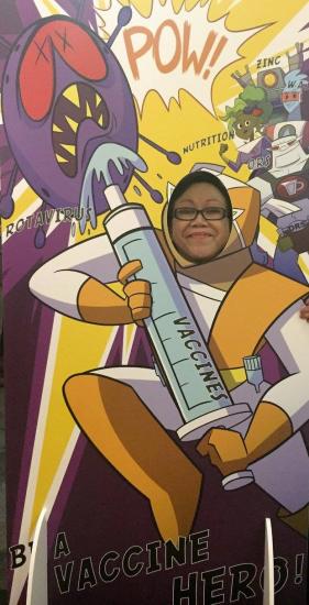 A person smiles while posing with their face in a cutout of an animated superhero character holding a giant syringe labeled "VACCINES." The background features comic book-style illustrations with words like "POW," a purple virus creature, and a cityscape.