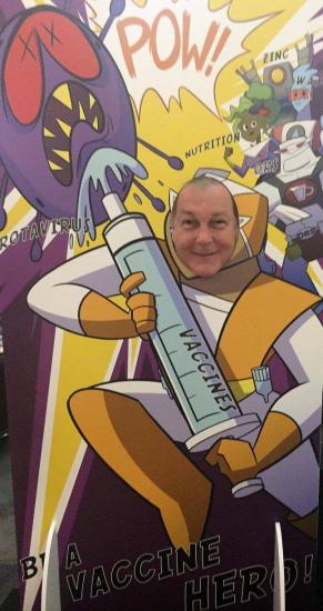 A person is posing with their face through a cutout of a cartoon superhero holding a giant syringe labeled "VACCINE" against a colorful backdrop featuring cartoon germs and the text "BE A VACCINE HERO!" on the bottom. The person appears to be smiling.