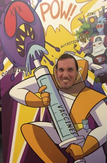 A man poses with his face in a cutout standee depicting a cartoon superhero holding a large syringe labeled "VACCINES". Above him, sound effect words like "POW!" and an illustrated virus monster labeled "ROTAVIRUS" add a dynamic, comic-book style backdrop.